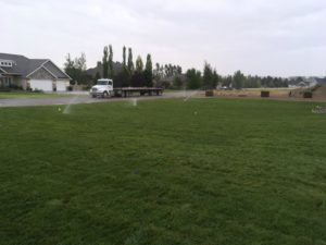 A new sod lawn being watered. The flatbed semi truck that delivered the sod is parked on the road in the background.